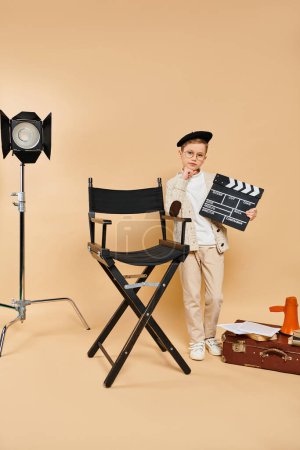 A young boy dressed as a film director holds a movie clapper next to a chair.