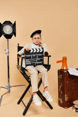 Preadolescent boy in director attire with movie clapper, seated in chair.