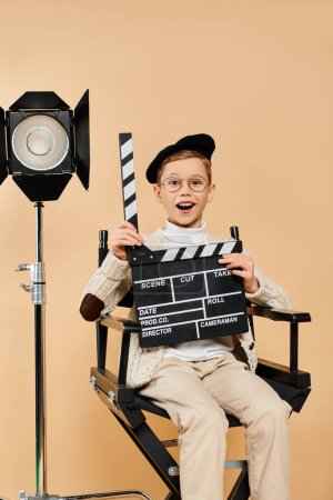 Vorpubertärer Junge posiert als Filmregisseur, sitzt im Stuhl und hält Filmklöppel in der Hand.