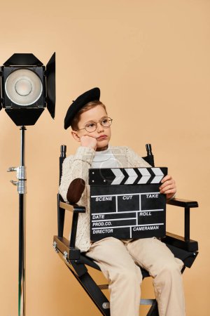 Als Filmregisseur verkleideter frühpubertärer Junge sitzt im Stuhl und hält Filmschiefer.