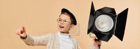 Photo for A young boy in glasses and a hat, holding a camera, dressed as a film director on beige backdrop. - Royalty Free Image