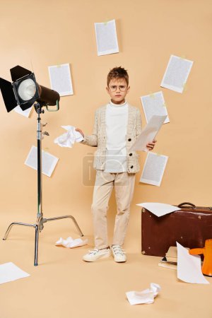 A cute preadolescent boy, dressed as a film director, stands confidently in front of the camera on a beige backdrop.