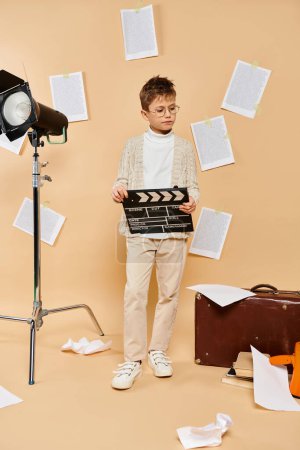 Preadolescent boy holds movie clapper in front of camera on beige backdrop.