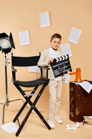A preadolescent boy in film director attire holding a movie clapper next to a chair.