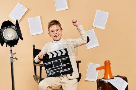 A preadolescent boy dressed as a film director sits with a movie clapper on a beige backdrop.