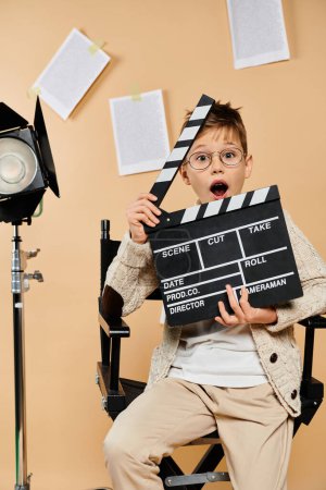 Young boy in film director attire, holding movie clapper in chair.