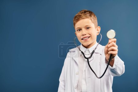 Preadolescent boy in doctors coat holds stethoscope against blue backdrop.