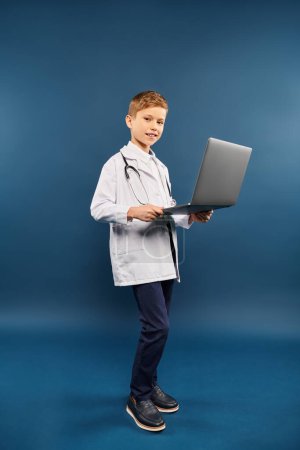 Un niño preadolescente con una bata de laboratorio sosteniendo un portátil contra un telón de fondo azul.