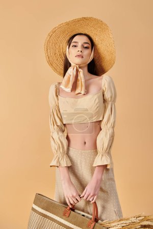A young woman with long brunette hair poses in a studio, dressed in a summer outfit, holding a bag and wearing a straw hat.