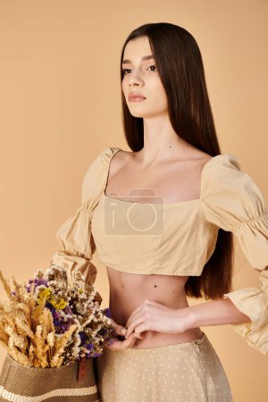 Une jeune femme aux longs cheveux bruns pose dans un crop top, tenant un bouquet vibrant de fleurs dans un décor studio.