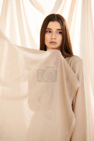 A young woman with long brunette hair strikes a pose in a summer outfit, exuding elegance and beauty in front of a white curtain.
