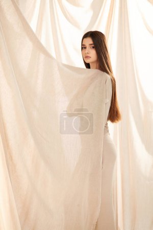 Eine junge Frau mit langen brünetten Haaren posiert vor einem weißen Vorhang und verbreitet in ihrem stylischen Outfit Sommerstimmung.