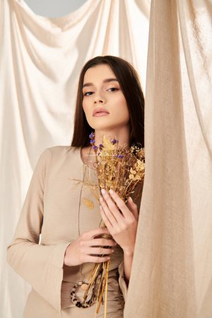 A young woman with long brunette hair holds a bunch of dried flowers, exuding a summery vibe in the studio.