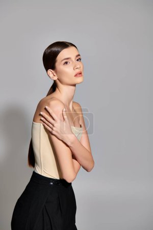 A young brunette woman poses confidently in a studio, showcasing a beige top and black pants on a grey background.