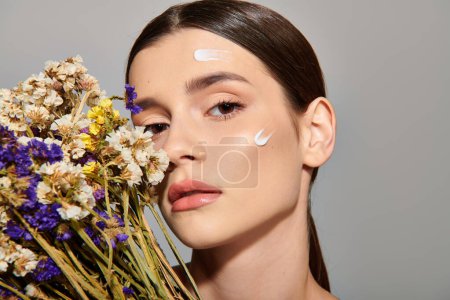 A young woman with brunette hair holds a bouquet of flowers while her face is painted white in a studio setting.