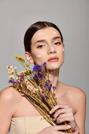 A brunette woman joyfully holds a bunch of flowers as bubbles float around her face in a studio setting on a grey background.