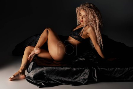 A woman in black lingerie sensually poses on a black sheet.