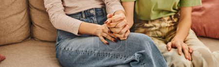 cropped banner of young lesbian couple holding hands and sitting on sofa, love and comfort at home