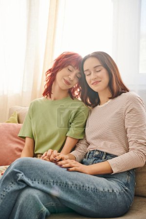 tenderness, young and pleased lesbian couple in casual attire holding hands and sitting on sofa