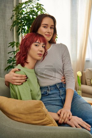 happy lesbian woman in 20s sitting on laps of her girlfriend while resting together in living room