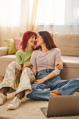 Foto de Serene moment of cheerful lesbian couple sitting near laptop and watching movie in living room - Imagen libre de derechos