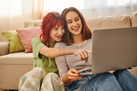 glücklicher Moment eines fröhlichen lesbischen Paares, das auf seinen Laptop zeigt und sich im Wohnzimmer einen Film ansieht