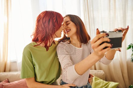 Photo for Young lesbian couple smiling and taking selfie on retro camera, capturing blissful moment at home - Royalty Free Image