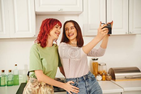 junges lesbisches Paar lächelt und macht Selfie mit Retro-Kamera in Küche und fängt glücklichen Moment ein