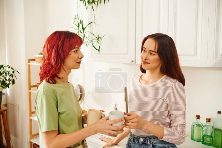 happy lesbian couple sharing a warm beverage while holding cups in their kitchen filled with light