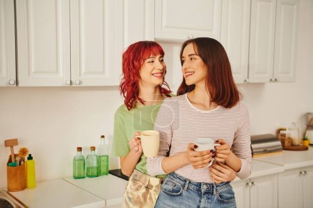happy lesbian couple sharing a warm beverage while holding cups in kitchen filled with light