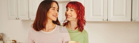 happy lesbian couple sharing a warm hug while standing in kitchen filled with light, banner