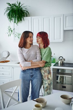 Foto de Happy and pierced lesbian woman with red hair embracing her girlfriend in modern kitchen - Imagen libre de derechos