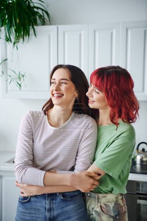 Foto de Retrato de alegre mujer lesbiana perforada con el pelo rojo mirando a su novia en la cocina - Imagen libre de derechos
