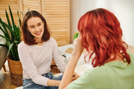 glückliche junge lesbische Frau schaut ihre Freundin mit roten Haaren an und lächelt im modernen Schlafzimmer