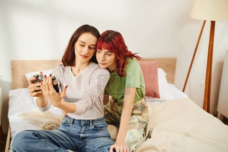 Foto de Candid casa sesión de fotos de jóvenes lesbianas pareja tomando selfie en retro cámara en el dormitorio - Imagen libre de derechos