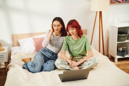 Téléchargez les photos : Heureux jeune couple lesbien dans casual tenue regarder film en ligne ensemble sur ordinateur portable dans chambre - en image libre de droit
