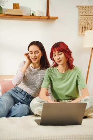 Téléchargez les photos : Sourire jeune couple lesbien en tenue décontractée regarder film en ligne ensemble sur ordinateur portable dans la chambre - en image libre de droit