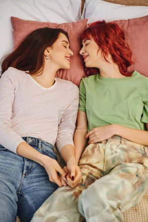 Téléchargez les photos : Top view of smiling lesbian couple holding hands and lying together on bed, looking at each other - en image libre de droit