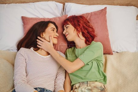 vista superior de la sonriente pareja de lesbianas mirándose y acostados juntos en la cama, tocando la cara