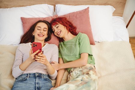 top view of happy young lesbian couple taking selfie on smartphone while lying together on bed
