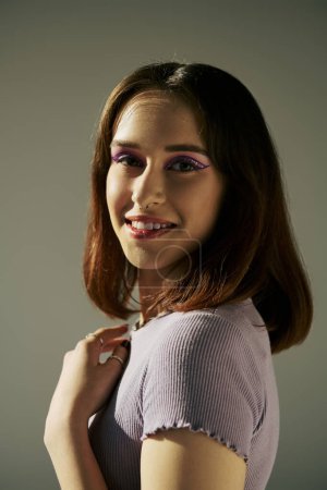 bold makeup of pleased gen z girl posing in t-shirt and looking at camera, purple eye shadow