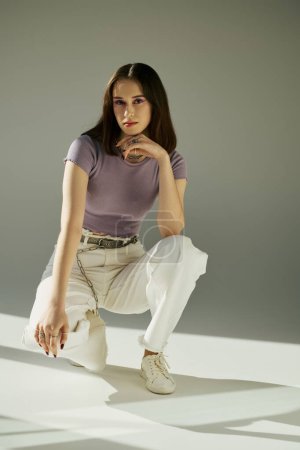 stylish young woman in purple t-shirt and white jeans with chain belt sitting on haunches on grey
