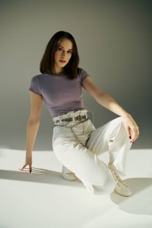 Jeune femme en t-shirt violet et jean blanc tendance avec ceinture à chaîne assise sur fond gris