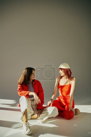 Téléchargez les photos : Heureux élégant couple lesbienne en tenue vibrante assis et regardant les uns les autres en studio sur gris - en image libre de droit