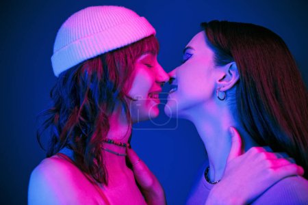 Foto de Beso sensual bajo luces moradas de alegre pareja lésbica con maquillaje audaz, generación z amor - Imagen libre de derechos
