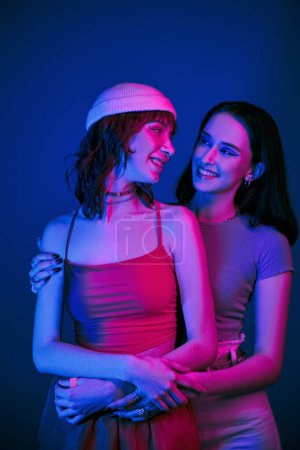 Foto de Tierno momento bajo luces púrpuras de alegre pareja lesbiana con maquillaje audaz, generación z amor - Imagen libre de derechos