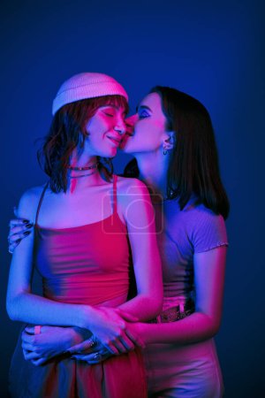 Foto de Beso sensual en la mejilla bajo luces púrpuras de feliz pareja lesbiana con maquillaje audaz, dicha y amor - Imagen libre de derechos