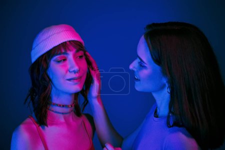 Zärtlicher Moment unter lila Lichtern einer lesbischen Frau mit kühnem Make-up, die das Gesicht ihrer Freundin berührt