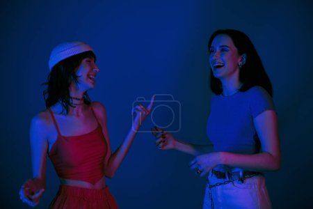 happy women in 20s, with bold makeup and stylish attire laughing and gesturing under purple lights