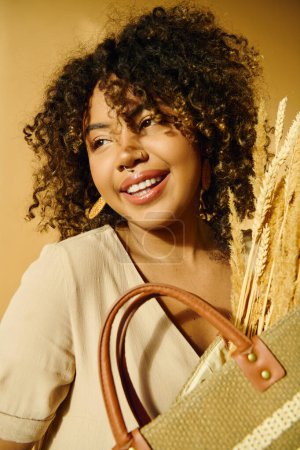 Foto de A beautiful young African American woman with curly hair smiling while holding a brown purse in a studio setting. - Imagen libre de derechos